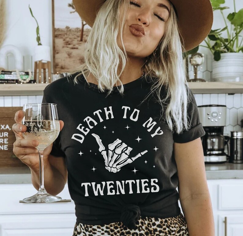May include: Black t-shirt with white text that reads "Death to my twenties" and a white skeleton hand making a shaka sign.