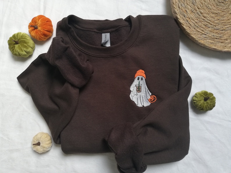Puede incluir: Una sudadera marr&oacute;n con un fantasma bordado que lleva un gorro naranja y sostiene una taza de caf&eacute;. El fantasma sostiene una peque&ntilde;a calabaza naranja en su otra mano.