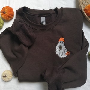 Puede incluir: Una sudadera marr&oacute;n con un fantasma blanco que sostiene una taza de caf&eacute; y lleva un gorro naranja. El fantasma est&aacute; bordado en la parte delantera de la sudadera. Hay una peque&ntilde;a calabaza naranja al lado del fantasma.