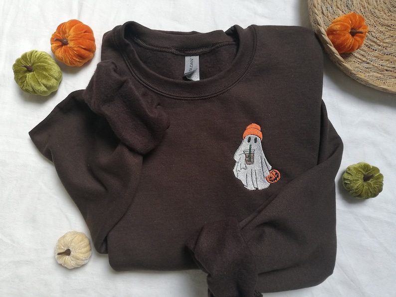 Puede incluir: Una sudadera marr&oacute;n con un fantasma blanco que sostiene una taza de caf&eacute; y lleva un gorro naranja. El fantasma est&aacute; bordado en la parte delantera de la sudadera. Hay una peque&ntilde;a calabaza naranja al lado del fantasma.