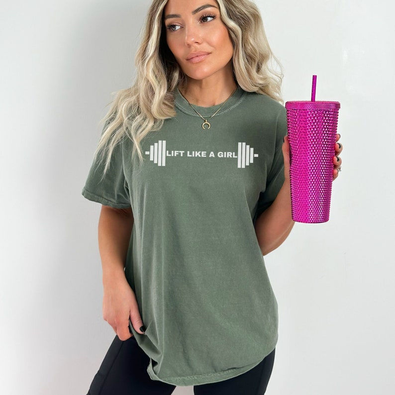 K&ouml;nnte beinhalten: Ein gr&uuml;nes T-Shirt mit dem Text "Lift Like A Girl" und einer wei&szlig;en Hantelgrafik. Die Person h&auml;lt einen rosa glitzernden Becher.