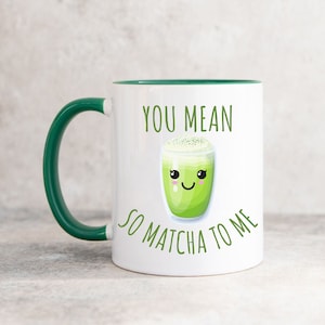 May include: A white ceramic mug with a dark green handle and interior rim. The front features a graphic of a smiling matcha latte in a glass with the green text "YOU MEAN SO MATCHA TO ME" printed above and below the illustration of the drink.