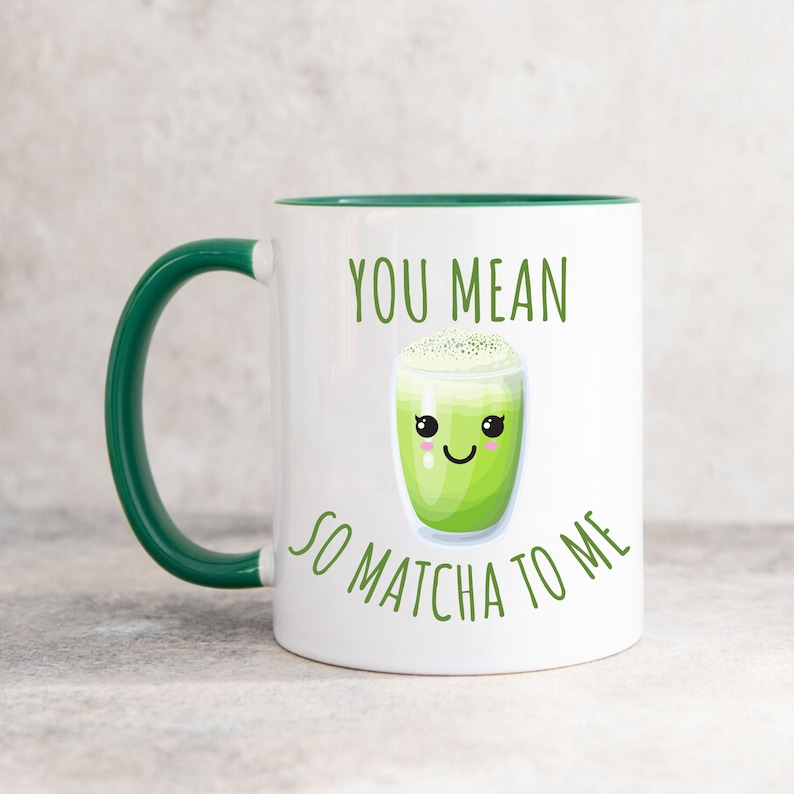 May include: A white ceramic mug with a dark green handle and interior rim. The front features a graphic of a smiling matcha latte in a glass with the green text "YOU MEAN SO MATCHA TO ME" printed above and below the illustration of the drink.