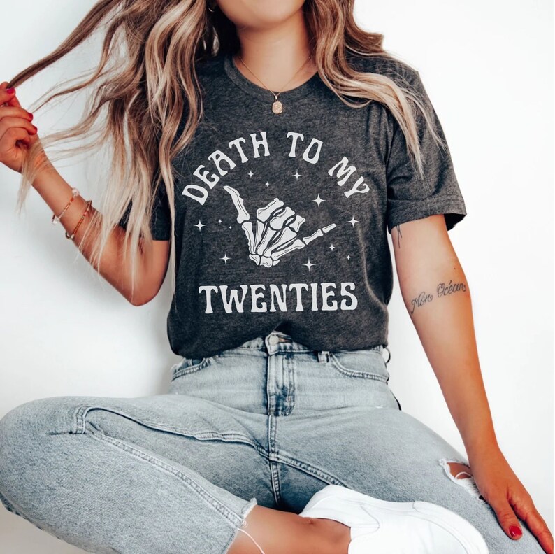 May include: A gray t-shirt with the text "Death to my twenties" and a skeleton hand making a shaka sign. The shirt has a white graphic print and is worn by a person with long blonde hair.