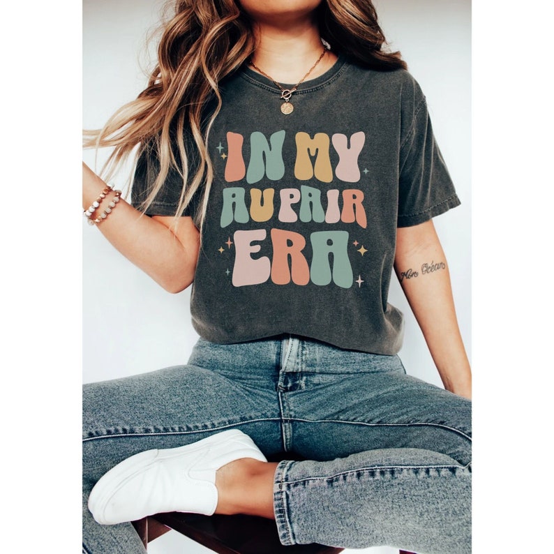May include: A dark grey t-shirt with the text "IN MY AUPAIR ERA" in colourful, retro-style lettering. The text is surrounded by small stars.