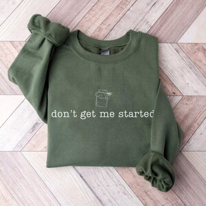 Könnte beinhalten: Ein olivgrünes Sweatshirt mit dem weißen Schriftzug "don't get me started" und einer kleinen Gläser-Illustration. Das Kleidungsstück liegt auf einer hellbraunen und weißen Holzoberfläche. Die Ärmel sind an den Handgelenken gerafft.