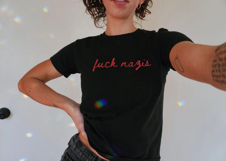 以下が含まれることがあります： 赤い筆記体で「fuck nazis」と書かれた黒いTシャツです。