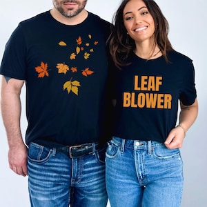 May include: Two black t-shirts with fall-themed designs. One shirt features orange and yellow leaves, while the other says "LEAF BLOWER" in orange letters. Both shirts are paired with blue jeans.