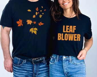 May include: Two black t-shirts with fall-themed designs. One shirt features orange and yellow leaves, while the other says "LEAF BLOWER" in orange letters. Both shirts are paired with blue jeans.