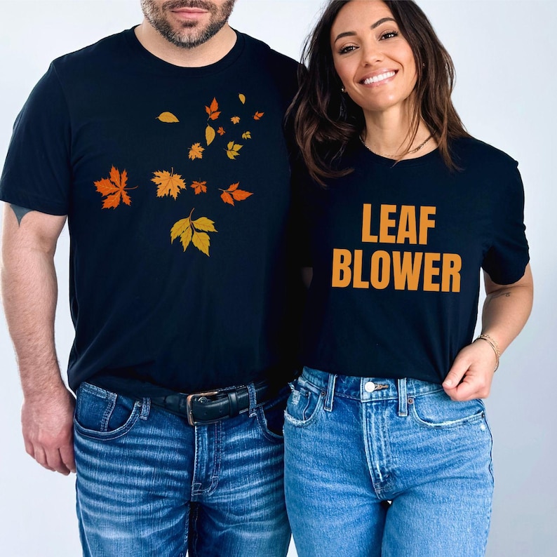 May include: Two black t-shirts with fall-themed designs. One shirt features orange and yellow leaves, while the other says "LEAF BLOWER" in orange letters. Both shirts are paired with blue jeans.