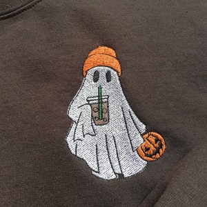 Puede incluir: Una sudadera marr&oacute;n con un dise&ntilde;o bordado de un fantasma que lleva una boina naranja y sostiene una taza de caf&eacute;. El fantasma sostiene una peque&ntilde;a calabaza naranja en su otra mano.