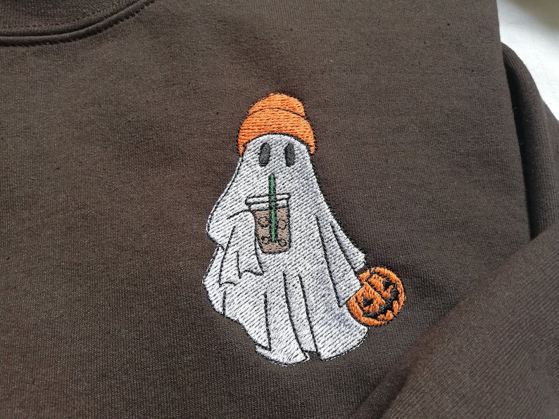 Puede incluir: Una sudadera marr&oacute;n con un dise&ntilde;o bordado de un fantasma que lleva una boina naranja y sostiene una taza de caf&eacute;. El fantasma sostiene una peque&ntilde;a calabaza naranja en su otra mano.