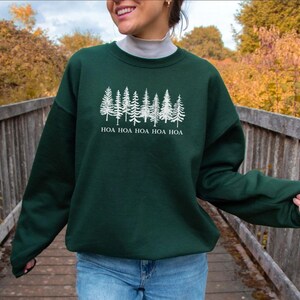 Peut inclure: Un sweat-shirt vert fonc&eacute; avec un motif blanc d'arbres de pins et le texte "HOA HOA HOA HOA HOA". Le sweat-shirt est &agrave; manches longues et &agrave; col rond. La personne sur l'image porte un jean bleu.