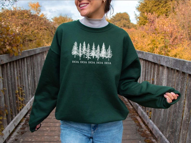 Peut inclure: Un sweat-shirt vert fonc&eacute; avec un motif blanc d'arbres de pins et le texte "HOA HOA HOA HOA HOA". Le sweat-shirt est &agrave; manches longues et &agrave; col rond. La personne sur l'image porte un jean bleu.