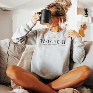May include: A light gray sweatshirt with the text "W+I+T+C+H WOMAN IN TOTAL CONTROL OF HERSELF" in black. The sweatshirt has long sleeves and a crew neck. A person is holding a black mug.