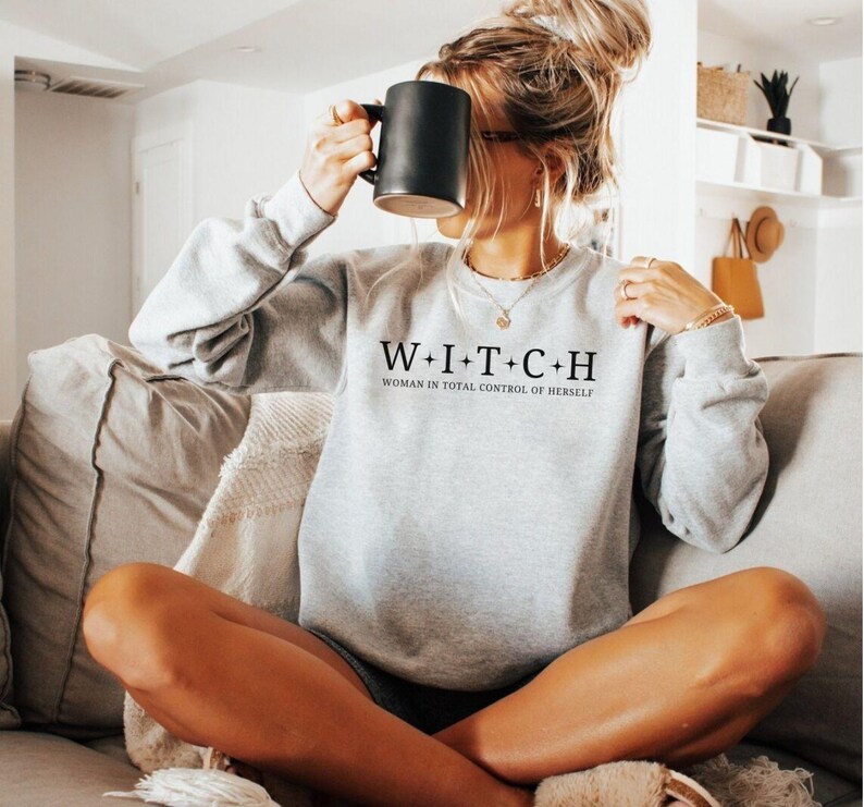 May include: A light gray sweatshirt with the text "W+I+T+C+H WOMAN IN TOTAL CONTROL OF HERSELF" in black. The sweatshirt has long sleeves and a crew neck. A person is holding a black mug.