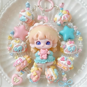 May include: A whimsical keychain featuring a doll-like figure holding a teddy bear, surrounded by pastel-colored charms. The charms include stars, hearts, and miniature cakes, all connected by beaded strands and a pink clasp. The color palette is soft and dreamy.