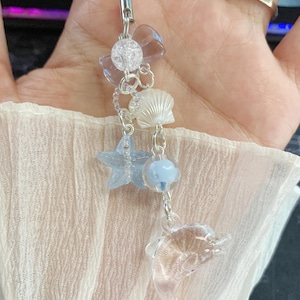 May include: A clear dolphin charm with a blue star, seashell, and bead accents. The charm is attached to a white cord with a silver clasp. The charm is a beach or ocean themed accessory.