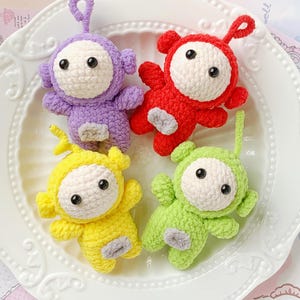 May include: Four handmade crochet dolls in purple, red, yellow, and green, each with a white face, black eyes, and a gray patch. The dolls are arranged on a white, scalloped plate. The dolls have a loop at the top.