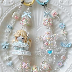 May include: Two keychains with pastel-colored charms. One keychain has a yellow ring and the other has a blue ring. Charms include a doll, star, flowers, bows, and heart-shaped beads. The keychains are on a white, decorative plate.