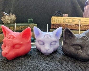 Puede incluir: Tres velas con forma de gato, cada una de un color diferente: rojo, morado y negro. Cada vela tiene un tercer ojo en la frente.