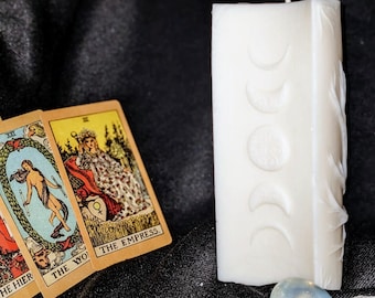 May include: A white, rectangular candle with a moon phase design and a wick. Tarot cards with the words "THE EMPRESS" are next to the candle. Several gemstones are at the base of the candle.