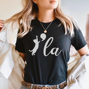 May include: Black t-shirt with a white silhouette of a cat reaching for a speech bubble with a comma inside. The text "'la" is written in white cursive below the cat.