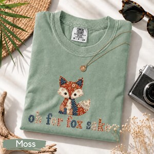 May include: A sage green t-shirt featuring a patchwork-style fox graphic and the text "Oh for fox sake" in a multi-colored floral and plaid pattern. The shirt has a crew neckline and a Comfort Colors brand label visible at the inner collar.