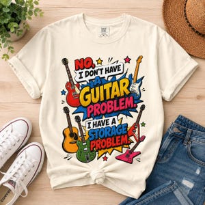 May include: A cream-colored short-sleeve t-shirt featuring a graphic print with five different guitars and the text "NO, I DON'T HAVE A GUITAR PROBLEM. I HAVE A STORAGE PROBLEM." The design includes red, yellow, blue, and green accents.