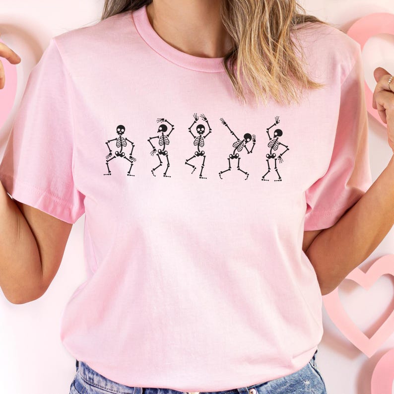 May include: Pink t-shirt with a graphic of five skeletons dancing. The skeletons are black and are in various poses.