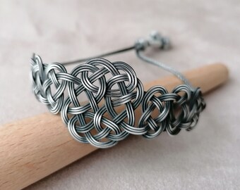 May include: A silver wire bracelet with a woven knot design. The bracelet is on a wooden surface.