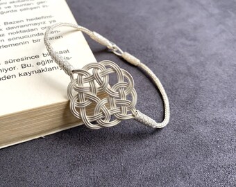 May include: A silver bracelet with a knotted design in the center. The bracelet is on a book.