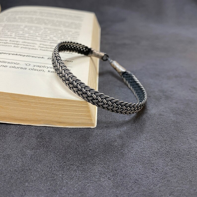 May include: A silver braided bracelet with a black leather backing. The bracelet is lying on a book.