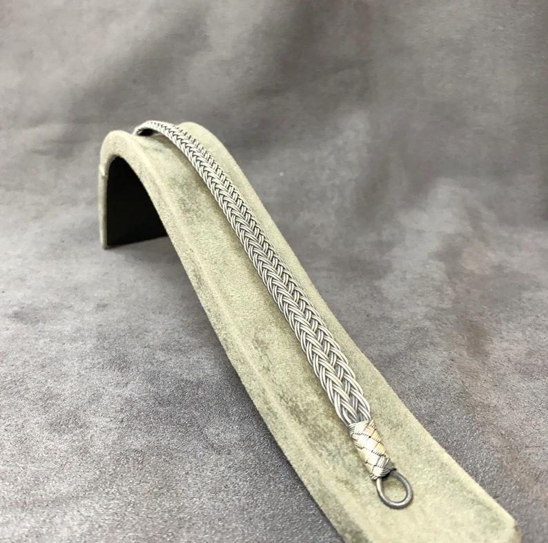 May include: A silver braided bracelet with a clasp. The bracelet is displayed on a gray fabric stand.
