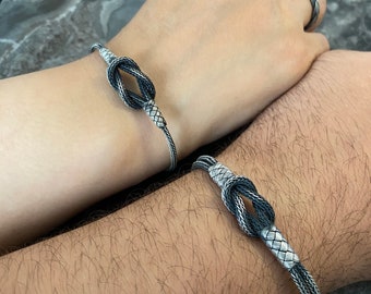 May include: Two silver bracelets with a knot design. The bracelets are made of braided silver wire with a black finish.