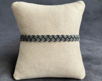 Op de afbeelding: Een zwarte gevlochten leren armband op een lichtbeige kussen.