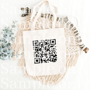May include: A white tote bag with a black QR code printed on the front. The bag is lying on a white surface with a woven macrame wall hanging and a branch behind it.