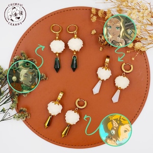 Könnte beinhalten: Eine Sammlung goldfarbener Ohrringe mit weißen, flauschigen Akzenten und Anhängern aus Edelsteinen oder Kristallen. Einige Ohrringe haben grüne, weiße oder goldfarbene Anhänger. Die Ohrringe werden auf einer braunen Lederoberfläche mit Illustrationen präsentiert.