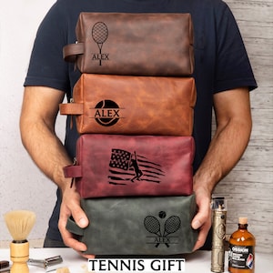 May include: Four leather toiletry bags in shades of brown, red, and green are stacked. Each bag features a tennis-themed design and the name "ALEX" or the text "TENNIS GIFT". The bags have a handle on the side.