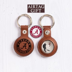 May include: Two brown leather AirTag keychains with silver rings. One keychain features the Alabama Crimson Tide logo, the other shows a coin with a bird design. The text "AIRTAG GIFT" is at the top.