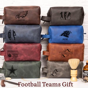 May include: A collection of leather toiletry bags in various colors, each featuring a football team logo and personalized name. The bags are rectangular with a handle and zipper closure. The image also includes a shaving brush, beard balm, and a straight razor.
