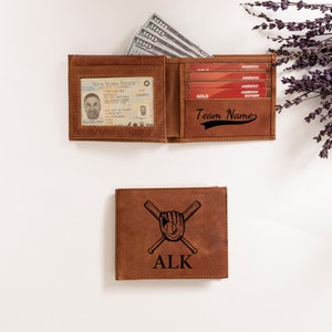 May include: Brown leather wallet with a baseball glove and crossed bats design. The wallet has the initials "ALK" engraved on the front. The inside of the wallet has a clear ID window and slots for credit cards. The wallet is open and has cash and credit cards inside.