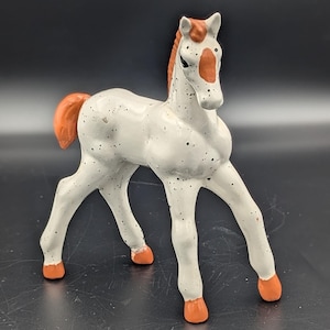 May include: A ceramic horse figurine featuring a white body with black speckles, a reddish-brown mane, tail, and hooves, and a reddish-brown patch on its forehead. The horse is standing in a forward-facing position with its head turned slightly.