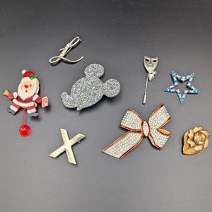 May include: A collection of eight assorted brooches including a Santa Claus figure, a silver-tone letter L, a glittery Mickey Mouse head, a theater mask stick pin, a blue rhinestone star, a gold-tone X, a rhinestone bow, and a floral brooch.