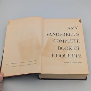 May include: Open book titled "Amy Vanderbilt's Complete Book of Etiquette." The book's cover is a light tan color with black text. The title is prominently displayed, and the book is open to a page with text and a taped area.