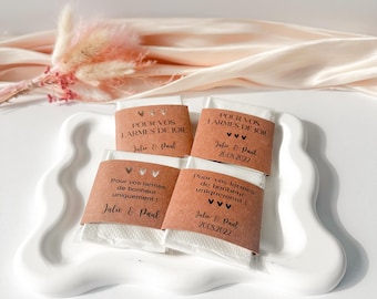 May include: Four small, wrapped wedding favors on a white, wavy-edged tray. Each favor has a brown paper label with the text "POUR VOS LARMES DE JOIE" and "Julie & Paul". The favors are for a wedding.