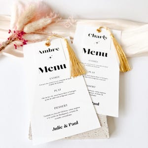 Può includere: Due biglietti bianchi per il menu di nozze con nappe dorate. Le carte hanno i nomi "Ambre" e "Charly" in alto, con la parola "Menu" in grassetto nero. Il menu include le opzioni antipasto, piatto principale e dessert. I nomi "Julie & Paul" sono in basso.