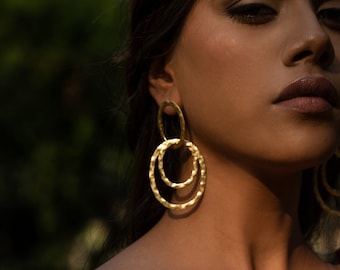 Pode incluir: Uma mulher usando brincos de argola de ouro com acabamento texturizado. Os brincos são de duas camadas e têm um visual martelado.