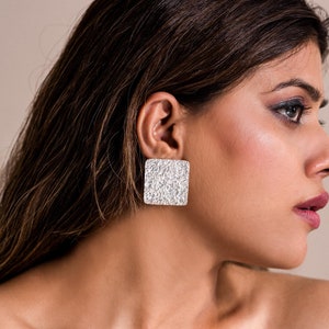 May include: A woman wearing silver square earrings with a textured surface. The earrings are large and eye-catching.