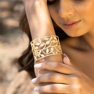 Peut inclure: Un bracelet manchette doré avec un motif de feuilles. Le bracelet entoure le poignet et présente un motif ajouré de feuilles et de vignes. Le bijou est une pièce de déclaration.